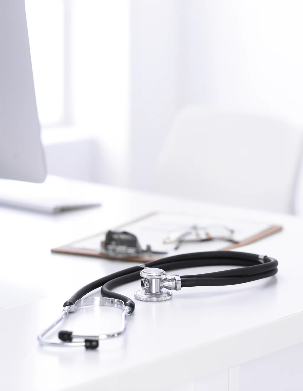 Stethoscope on a white desk, with a computer and clipboard in the background