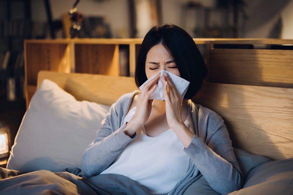 Woman sitting cozily on bed, holding a tissue to her nose seemingly blowing on it