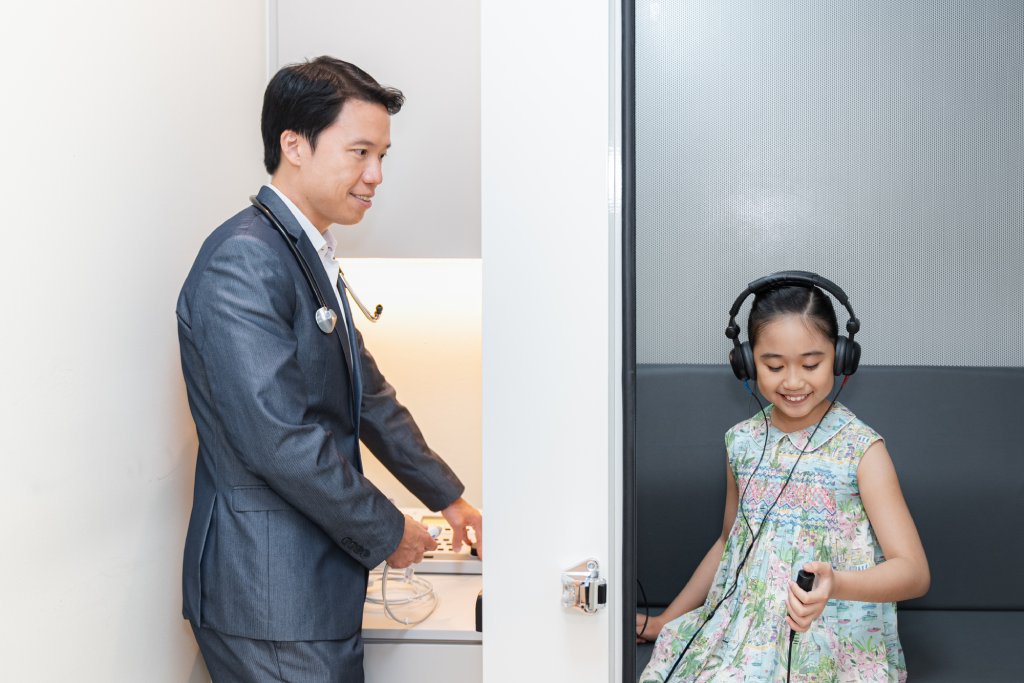 Dr. Chua in a suit adjusts equipment while a young girl in a dress wears headphones, sitting in a soundproof booth