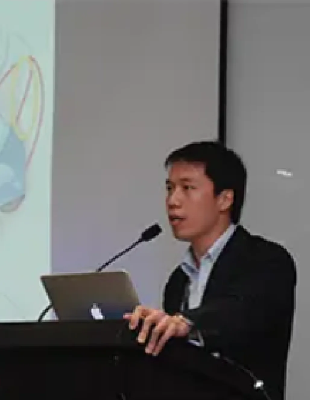 Dr. Chua stands at a podium, presenting to an audience with a laptop and a colorful diagram projected behind him